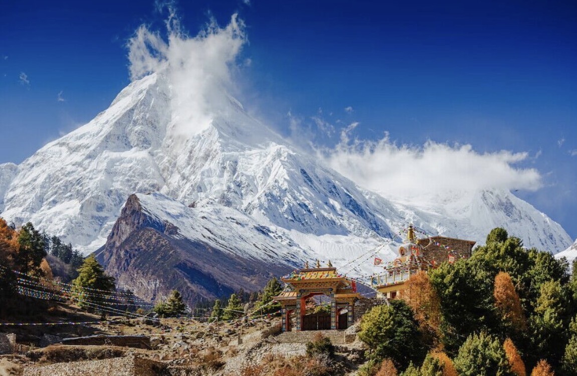 Snow-covered Himalayan peak rising above a hillside monastery and prayer flags in Nepal